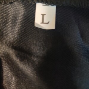 May include: Close-up of a black fabric with a white label. The label has the letter "L" printed on it, indicating a large size. The fabric appears to be a dark denim or similar material.