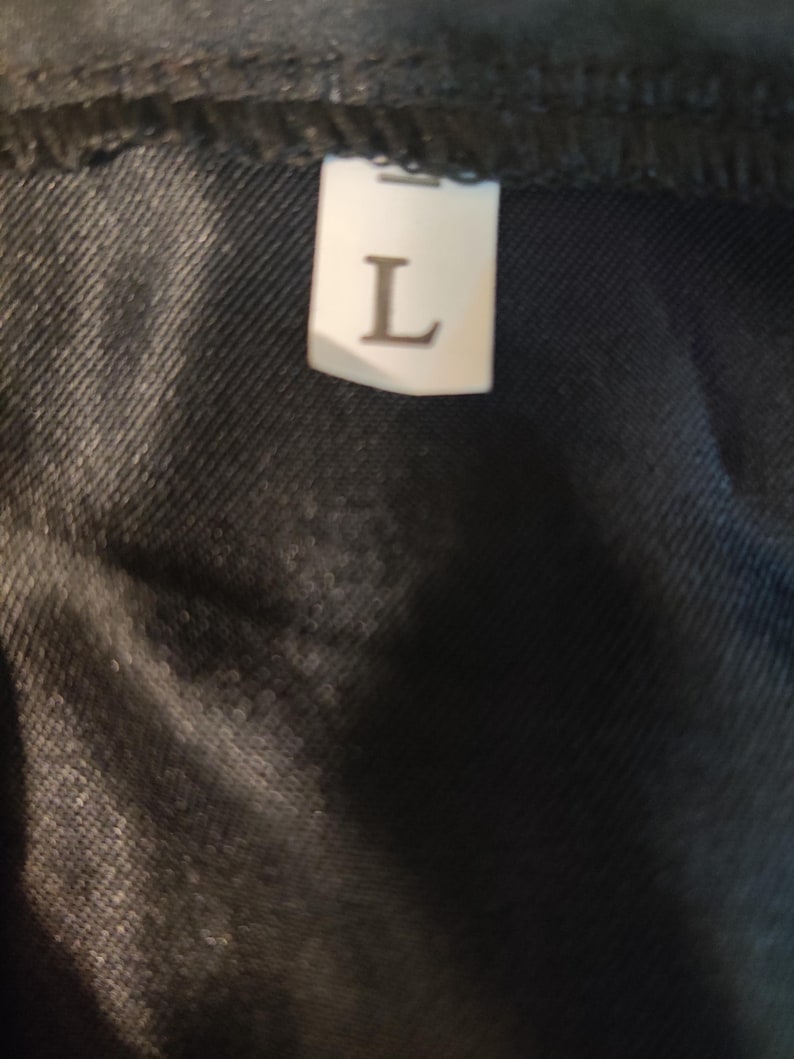 May include: Close-up of a black fabric with a white label. The label has the letter "L" printed on it, indicating a large size. The fabric appears to be a dark denim or similar material.