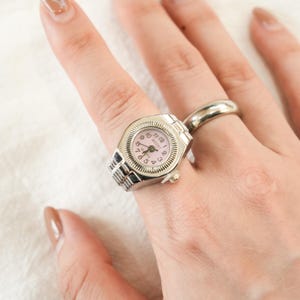 Op de afbeelding: Zilverkleurige horlogering met een roze wijzerplaat. De wijzerplaat toont de cijfers 1 tot en met 12 en de merknaam "BOZHI". De ring wordt aan een vinger gedragen, met een andere zilveren ring zichtbaar op de achtergrond.