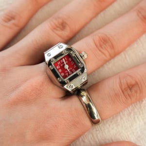May include: A silver ring featuring a watch face. The watch face is red with white numerals and hands. The watch has a small crown. The ring is worn on a finger.