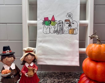 以下が含まれることがあります： ワッフル織りの白いキッチンタオル。ハロウィンの仮装をした5人のキャラクターのグラフィックがプリントされています。魔女2人と幽霊3人が描かれており、1人の幽霊が茶色の袋を持ち、I GOT A ROCKと書かれた吹き出しが付いています。
