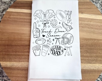 May include: A white kitchen towel featuring black line art illustrations of football-themed items including a helmet, football, shoulder pads, a foam finger, and a drink cup, with the text Touch Down Season and GAME DAY printed in the center.