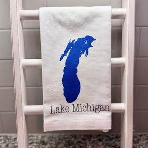 May include: A white waffle-weave kitchen towel featuring a blue graphic silhouette of Lake Michigan centered on the fabric. Below the blue lake shape, the text Lake Michigan is printed in a simple black serif font on the lower portion of the towel.