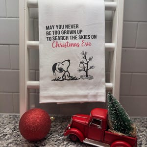 May include: A white waffle-weave kitchen towel featuring a black graphic of Snoopy wearing a Santa hat next to a small Christmas tree. The text reads: MAY YOU NEVER BE TOO GROWN UP TO SEARCH THE SKIES ON Christmas Eve, with the last two words in red.