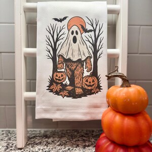 May include: A white Halloween-themed kitchen towel with a Bigfoot-ghost hybrid design. The illustration features a ghost-like figure with a Bigfoot body, holding a jack-o'-lantern. The background includes a full orange moon, bats, and bare trees. The towel is displayed on a white ladder.