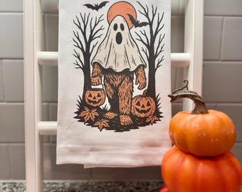May include: A white Halloween-themed kitchen towel with a Bigfoot-ghost hybrid design. The illustration features a ghost-like figure with a Bigfoot body, holding a jack-o'-lantern. The background includes a full orange moon, bats, and bare trees. The towel is displayed on a white ladder.