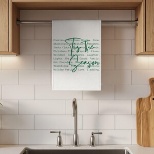 May include: A white kitchen towel hanging on a metal bar, featuring a list of holiday-themed words in green text and the phrase 'Tis the season' written in a larger, cursive green script across the center of the printed word list.