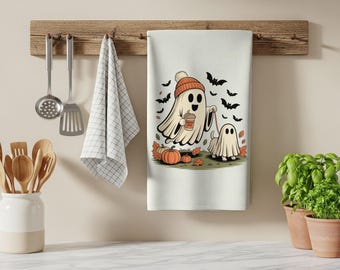 May include: A white kitchen towel featuring a graphic of a ghost wearing an orange beanie, holding a coffee cup, and walking a smaller ghost-dog on a leash. The scene includes orange pumpkins, falling autumn leaves, and black bats in the background.