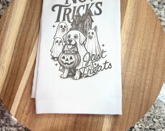 May include: A white kitchen towel featuring a dark gray graphic illustration of three dogs dressed as ghosts and one dog holding a pumpkin candy bucket. The text reads No TRICKS Just Treats, with a haunted house silhouette in the background.