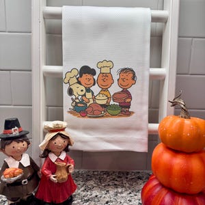 May include: White kitchen towel with a Thanksgiving-themed illustration featuring cartoon characters. The towel is hanging on a white wooden rack. Decorative figurines and stacked pumpkins are in the foreground.
