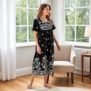 May include: A black, short-sleeved nightgown with a V-neck and floral embroidery. The dress features white and gray floral patterns along the hem, bodice, and sleeves. The model is standing in a well-lit room with a window and a chair.