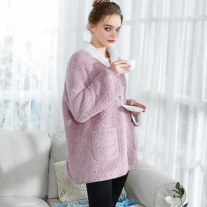 May include: A light purple, fuzzy robe with a white collar and pockets. The robe is worn over black leggings. The person is holding a white teacup and saucer. The background includes a white couch and curtains.