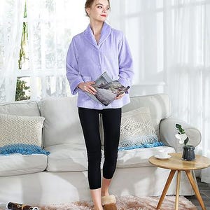 May include: A light purple, long-sleeved, textured robe with a shawl collar and a single button closure. The robe is paired with black leggings and tan, fuzzy slip-on slippers. The garment features a ribbed fabric texture throughout.