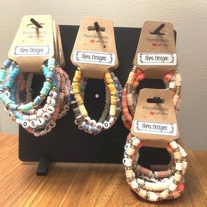 May include: Handmade bracelets in various colors, including blue, pink, and orange, with gold-colored accents. Some bracelets feature letter beads spelling out words. Each bracelet is displayed on a black stand with a tag that reads "Alira Designs."