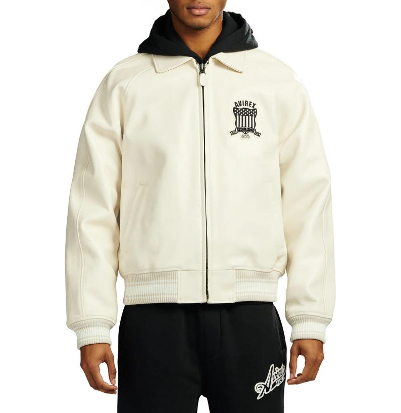 May include: Cream-colored jacket with a black hood and collar. The jacket features a zipper closure, ribbed cuffs, and a ribbed hem. The left chest displays a black emblem with the text "AVIREX" and "1975".