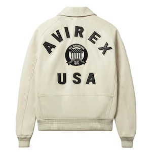 May include: Cream-colored leather jacket with a collar and ribbed cuffs. The back features the word "AVIREX" in large black letters above a circular emblem and the word "USA" below.