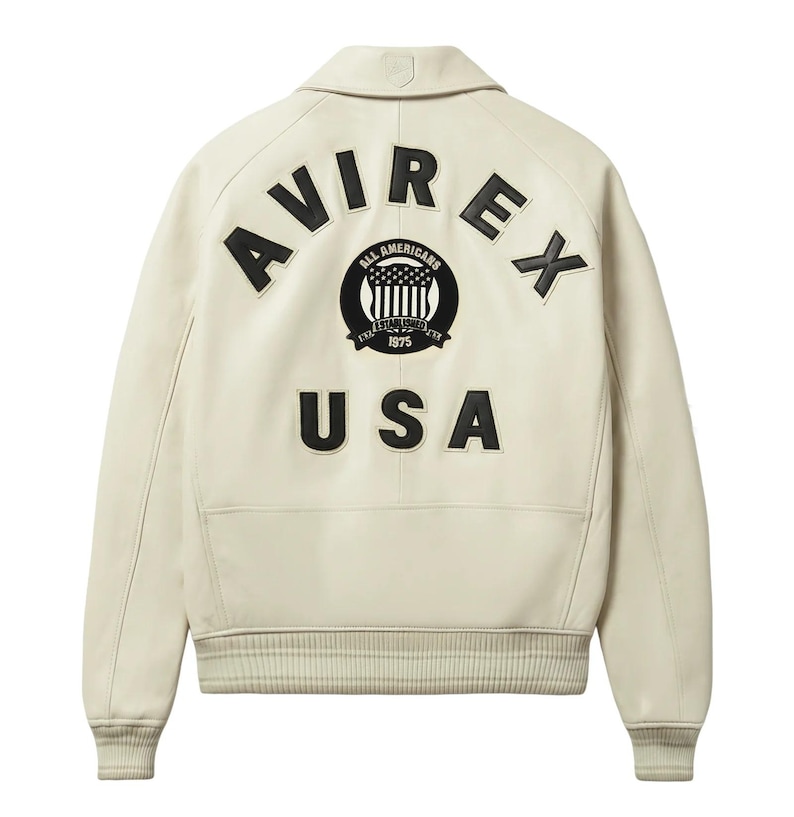 May include: Cream-colored leather jacket with a collar and ribbed cuffs. The back features the word "AVIREX" in large black letters above a circular emblem and the word "USA" below.