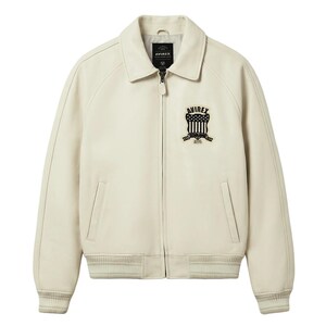 May include: Cream-colored leather jacket with a collared neckline, a zipper closure, and ribbed cuffs and hem. Features a black and white embroidered patch on the chest with the text "AVIREX" and a shield design. Includes two front pockets.