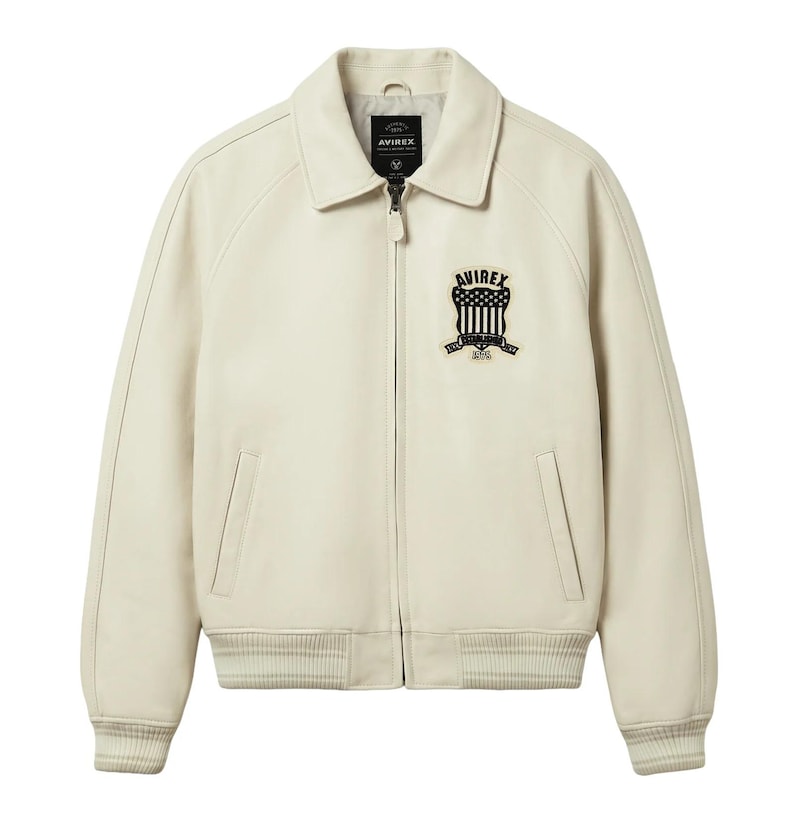 May include: Cream-colored leather jacket with a collared neckline, a zipper closure, and ribbed cuffs and hem. Features a black and white embroidered patch on the chest with the text "AVIREX" and a shield design. Includes two front pockets.