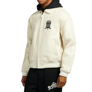 May include: Cream-colored bomber jacket with a black hood and collar. The jacket features a zipper closure, side pockets, and ribbed cuffs and hem. A black and white logo is on the chest. The model is wearing black sweatpants with a white logo.