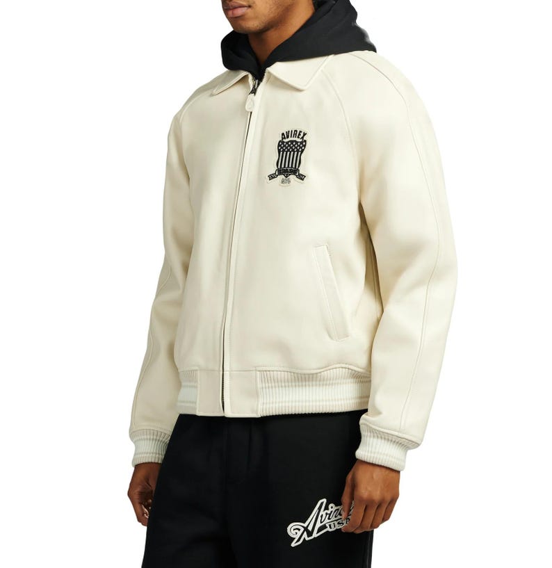 May include: Cream-colored bomber jacket with a black hood and collar. The jacket features a zipper closure, side pockets, and ribbed cuffs and hem. A black and white logo is on the chest. The model is wearing black sweatpants with a white logo.