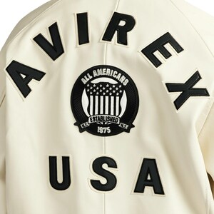 May include: Cream-colored jacket with black lettering and patches. The word "AVIREX" is across the top, and "USA" is at the bottom. A circular patch in the center reads "ALL AMERICANS ESTABLISHED 1975 N.Y."