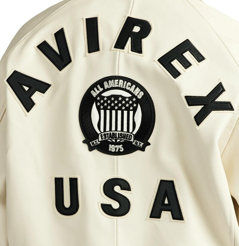 May include: Cream-colored jacket with black lettering and patches. The word "AVIREX" is across the top, and "USA" is at the bottom. A circular patch in the center reads "ALL AMERICANS ESTABLISHED 1975 N.Y."
