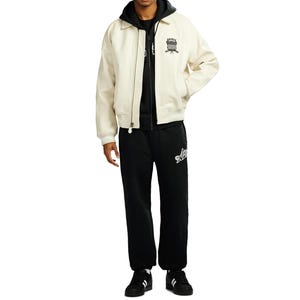 May include: Cream-colored bomber jacket with a black hoodie underneath, paired with black sweatpants. The jacket features a black emblem on the chest. The sweatpants have a white logo on the left leg. Black and white sneakers complete the outfit.