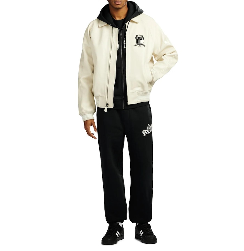 May include: Cream-colored bomber jacket with a black hoodie underneath, paired with black sweatpants. The jacket features a black emblem on the chest. The sweatpants have a white logo on the left leg. Black and white sneakers complete the outfit.