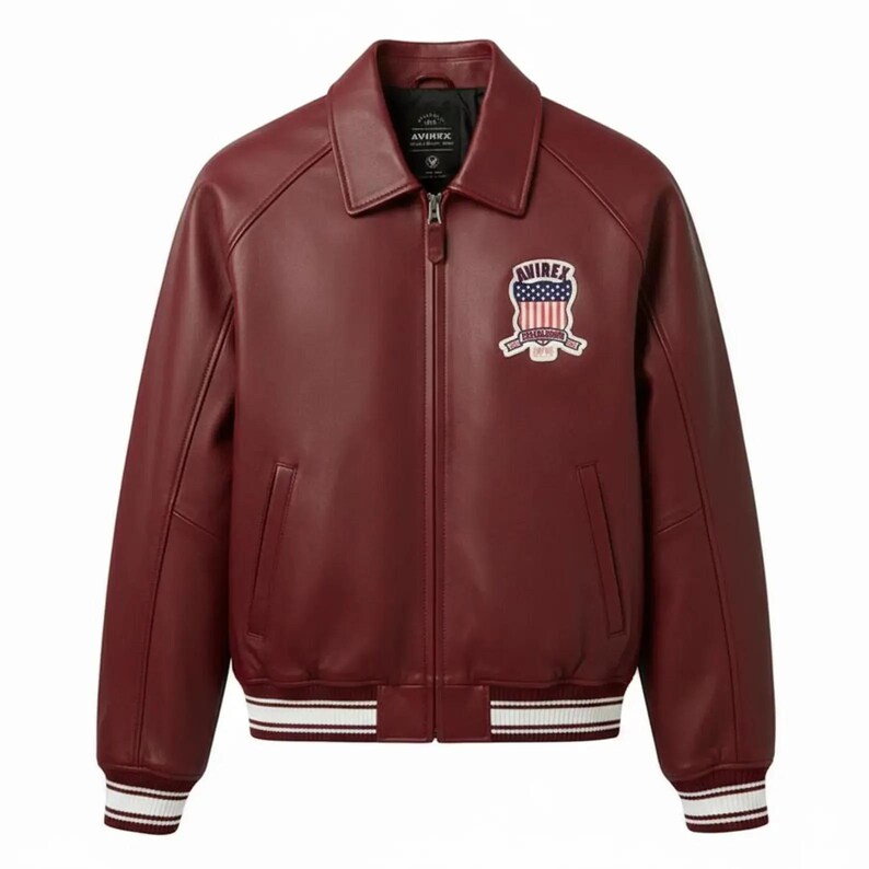 May include: A deep burgundy leather jacket featuring a classic collar, a full-length front zipper, and ribbed cuffs and hem with white stripes. A patch on the left chest displays the text AVIREX above a shield with the American flag design.