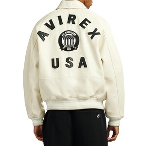 May include: Cream-colored leather jacket with the word "AVIREX" arched across the shoulders in black, along with a circular emblem and the word "USA". The jacket has a collared neckline and ribbed cuffs.