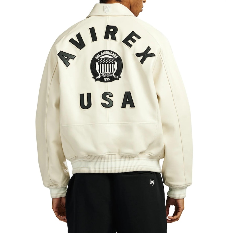May include: Cream-colored leather jacket with the word "AVIREX" arched across the shoulders in black, along with a circular emblem and the word "USA". The jacket has a collared neckline and ribbed cuffs.