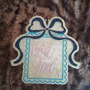 May include: A beige, rectangular hanging ornament with a scalloped edge and a large bow at the top. The bow is outlined in black glitter. The center features the text "But God" in pink glitter script, with a small pink glitter cross below.