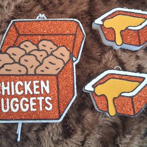 May include: A set of three glittery orange ornaments featuring a box labeled CHICKEN NUGGETS filled with tan nuggets and two smaller square containers filled with yellow dipping sauce, each with a white outline and a small metal hanging loop.