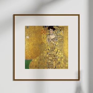 Puede incluir: Una impresión enmarcada de una pintura de Gustav Klimt, que presenta a una mujer con un vestido dorado adornado. La obra de arte se exhibe en una pared blanca, con un jarrón blanco y una planta verde en primer plano. El marco es marrón.