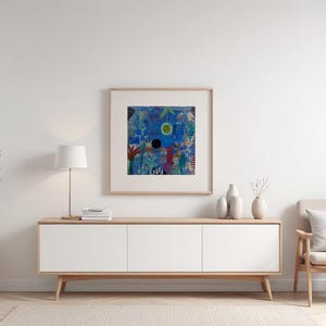 May include: An abstract painting in a wooden frame hangs above a white cabinet. The artwork features a blue background with colourful shapes and a central black circle. A beige chair and decorative vases complete the scene.