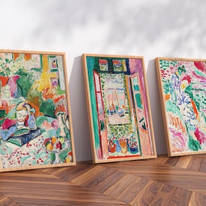 May include: Three framed art prints with colorful abstract paintings. The artwork features vibrant hues of green, red, blue, and yellow. The frames are made of light-colored wood, and the paintings lean against a white wall on a wooden floor.