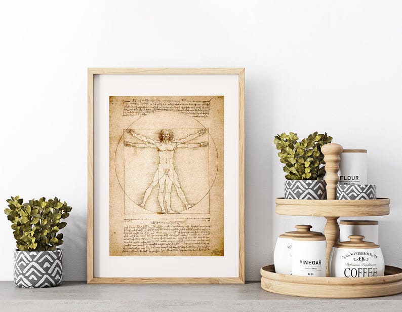 May include: A framed print of Leonardo da Vinci's Vitruvian Man. The artwork features a detailed sketch of a nude male figure within a circle and square, with handwritten notes. The frame is light wood, and the print has a sepia tone.