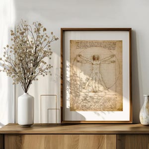 May include: A framed print of Leonardo da Vinci's Vitruvian Man, displayed on a wooden cabinet. The artwork features a detailed sketch of a man within a circle and square, with handwritten notes. A white vase with dried flowers and a decorative vase are also on the cabinet.