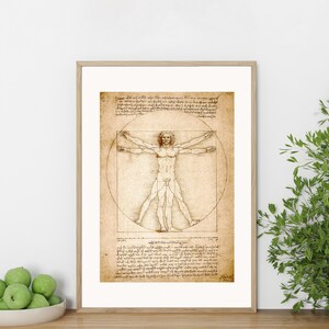 May include: Framed print of Leonardo da Vinci's Vitruvian Man. The artwork features a detailed pen and ink drawing of a nude male figure within a circle and a square, with handwritten text. The frame is a light wood color.
