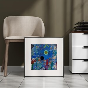 May include: Framed abstract art print with a vibrant blue background, featuring a central black circle and a yellow-green sun-like shape. The artwork includes red, green, and white botanical elements. The print is displayed near a beige chair and a white cabinet.