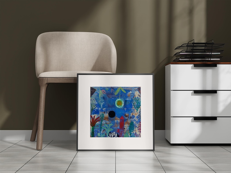 May include: Framed abstract art print with a vibrant blue background, featuring a central black circle and a yellow-green sun-like shape. The artwork includes red, green, and white botanical elements. The print is displayed near a beige chair and a white cabinet.