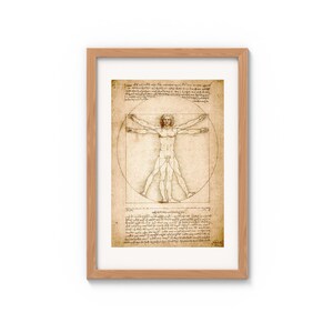 May include: A framed print of Leonardo da Vinci's Vitruvian Man. The artwork features a detailed pen and ink drawing of a nude male figure within a circle and square, with handwritten notes in the background. The frame is made of light brown wood.