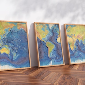 May include: Three framed relief maps of the world's ocean floors. The artwork features a detailed depiction of the ocean depths in shades of blue, yellow, and brown, set within wooden frames. The maps are leaning against a white wall.