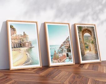 May include: Three framed art prints featuring coastal scenes. The prints depict Italian architecture, including a seaside town, a coastal view, and an arched stone doorway. The frames are light brown, and the images have a bright, sunny aesthetic.