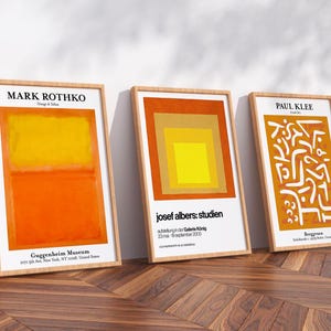 May include: Three framed art prints leaning against a white wall on a wooden floor. The prints feature abstract art by Mark Rothko, Josef Albers, and Paul Klee. The Mark Rothko print has orange and yellow blocks. The Josef Albers print has a square design. The Paul Klee print has white shapes.