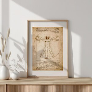 May include: Framed print of Leonardo da Vinci's Vitruvian Man. The artwork features a detailed sketch of a human figure within a circle and square, with handwritten notes. The frame is a light wood color, and the print has a vintage, aged appearance.