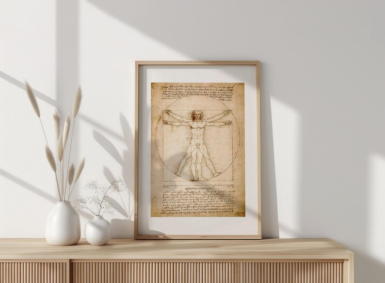 May include: Framed print of Leonardo da Vinci's Vitruvian Man. The artwork features a detailed sketch of a human figure within a circle and square, with handwritten notes. The frame is a light wood color, and the print has a vintage, aged appearance.