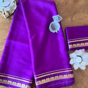 May include: A vibrant purple silk saree featuring a gold border with a repeating elephant motif. The fabric has a smooth, satin-like finish. A small silver-toned decorative container and two white lotus-shaped candle holders are placed nearby.