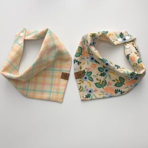 May include: Two dog bandanas. One is pastel plaid with pink, yellow, and blue. The other is cream-coloured with a floral pattern of blue, peach, and green. Both have snap closures and small brown tags.